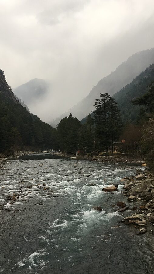 The Parvati river flowing through Kasol with stone banks and forested hills.