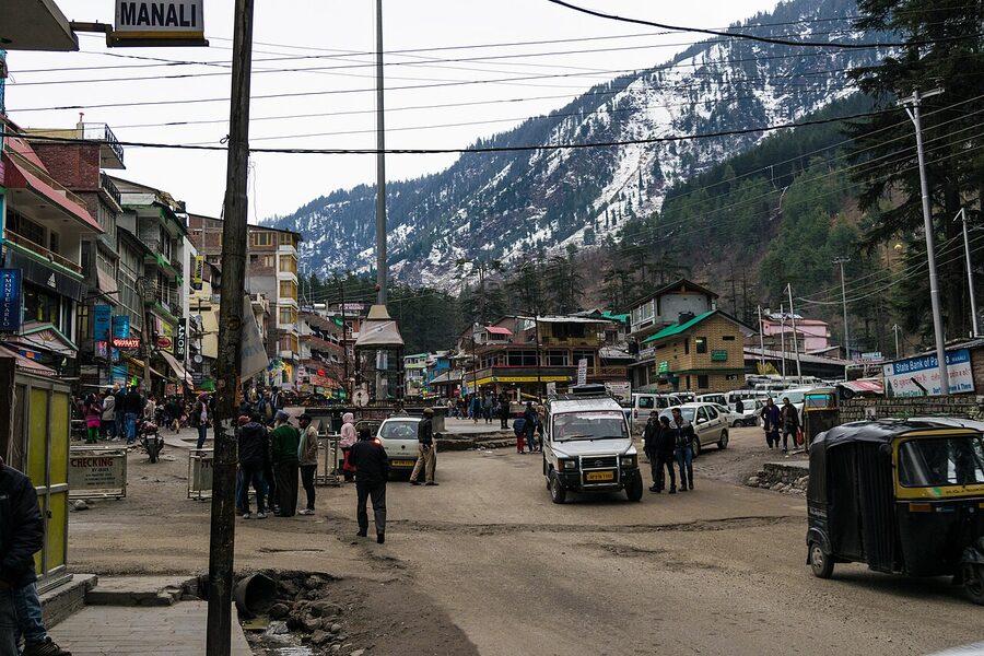 Shopfronts and signage along Manali Mall Road with the surrounding deodar slopes behind.